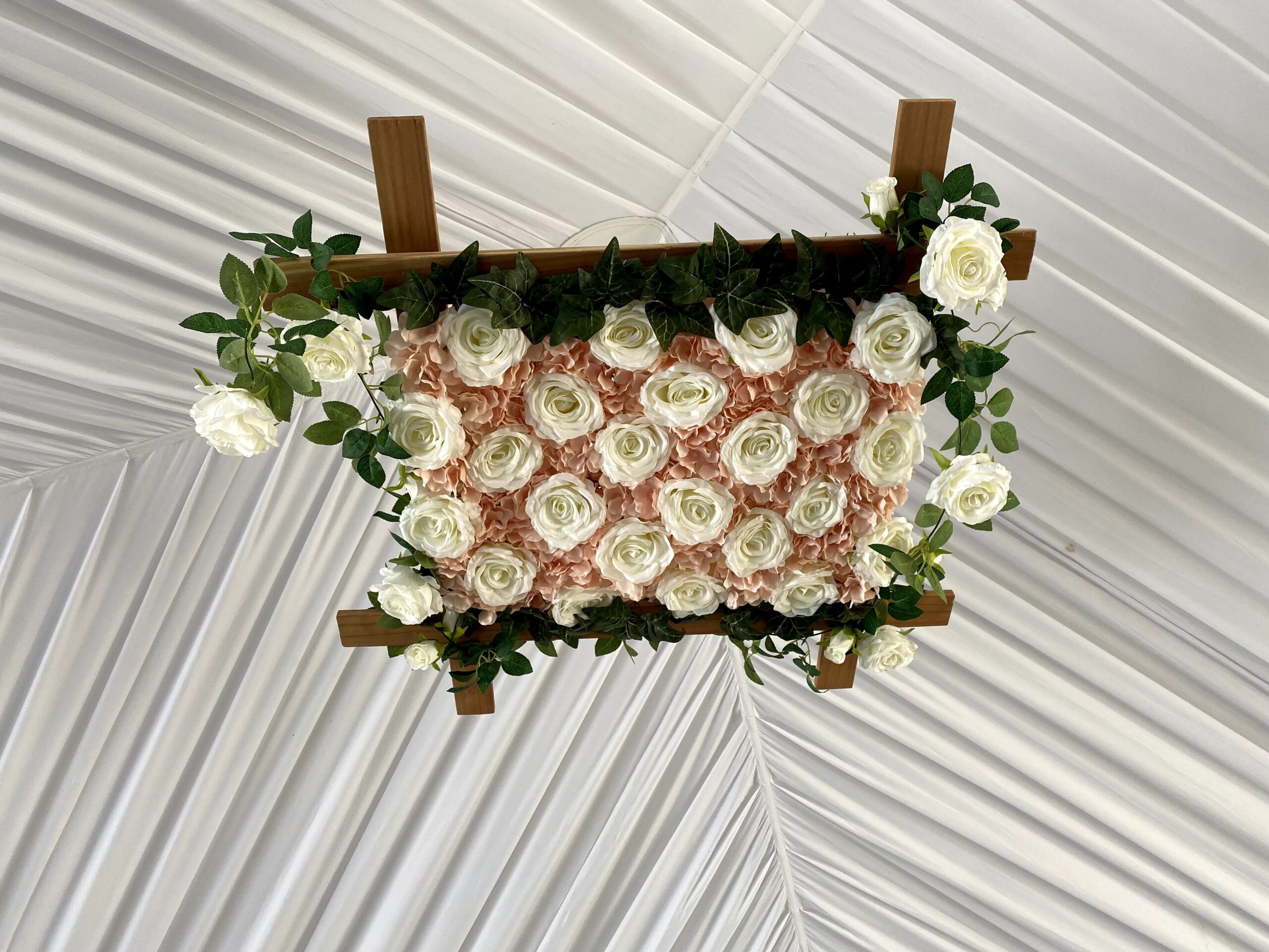 Rectangular flower arrangement with white and peach roses framed by wooden beams and green leaves.